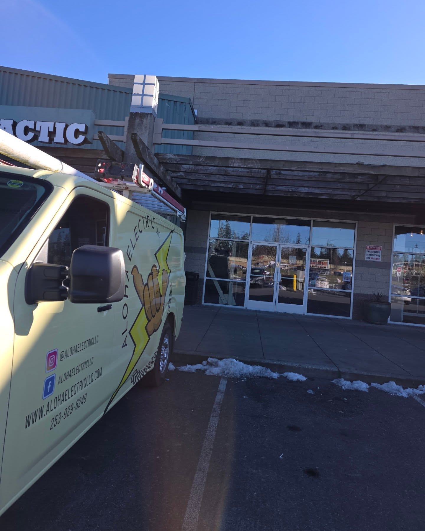 A yellow Aloha Electric van is parked outside a commercial building with glass doors and a sign above, highlighting their expertise in commercial electrical services. Snow is on the ground near the entrance.