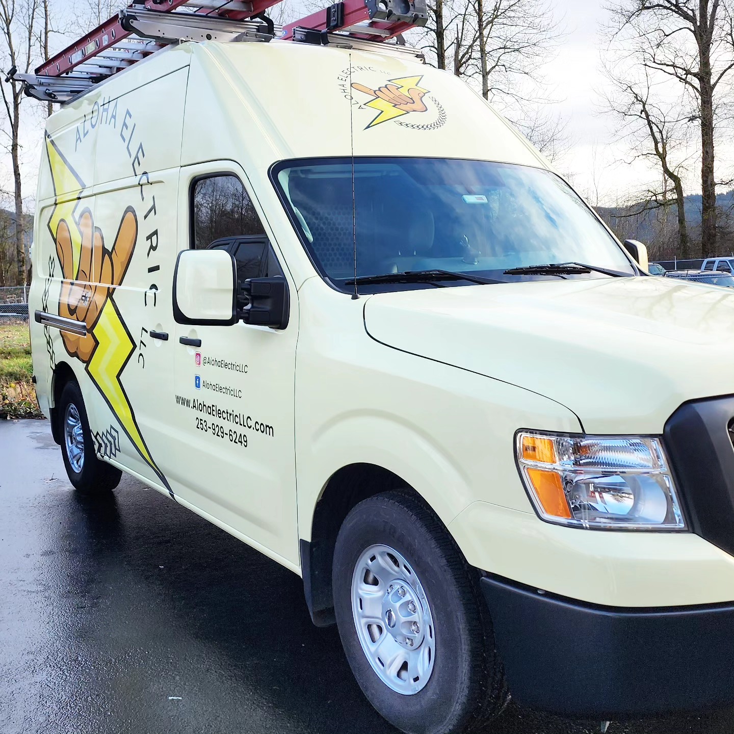 A yellow Aloha Electric Inc. van with company logo and contact details, specializing in residential electrical services, is parked on a wet pavement near leafless trees.