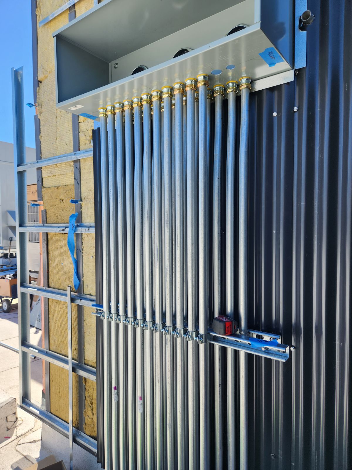 A row of metal electrical conduits is installed vertically on a corrugated wall, connected to a junction box at the top, with construction materials visible in the background.