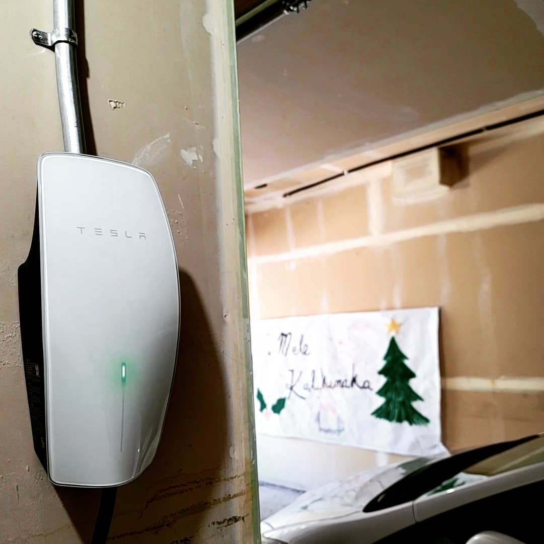 A Tesla wall charger is mounted on a garage wall with a car parked below and a Christmas-themed sign reading Mele Kalikimaka in the background.