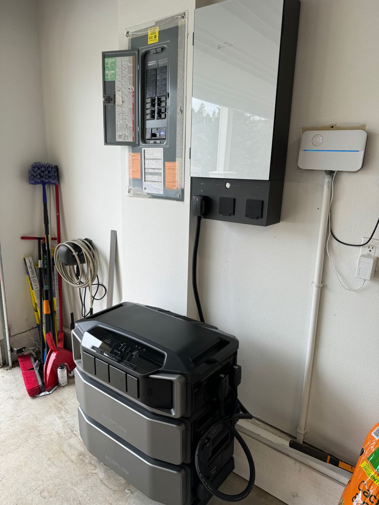 A home energy storage system with two battery units stacked below a wall-mounted inverter, installed next to an electrical panel in a garage. Cleaning tools are visible on the left.
