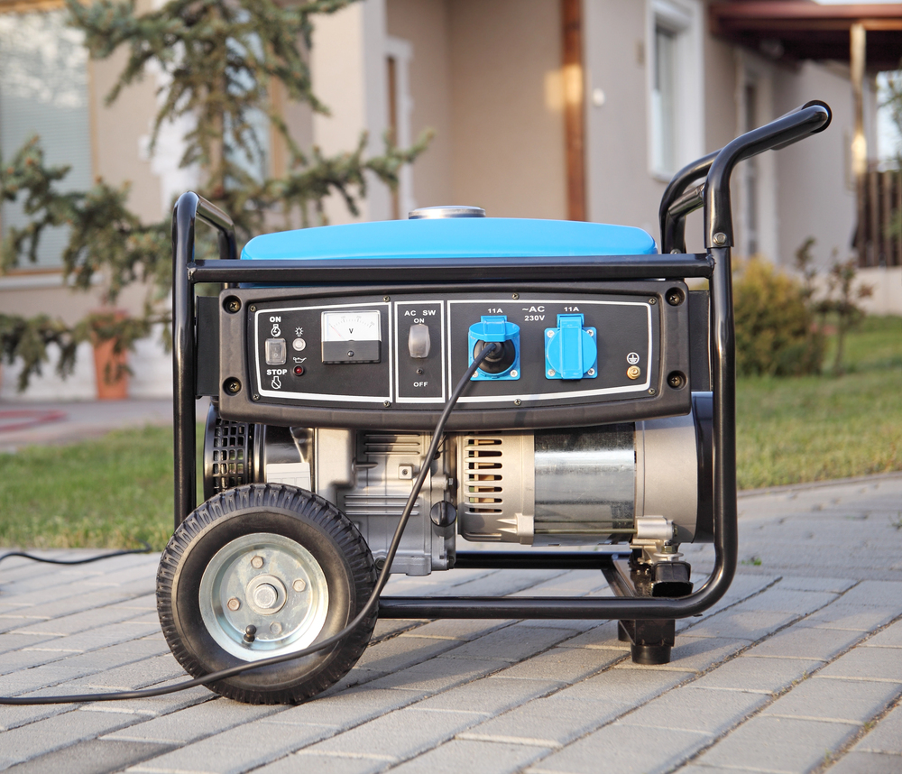 A portable gasoline generator with a blue top is placed on a paved walkway outdoors, with a house and trees in the background.