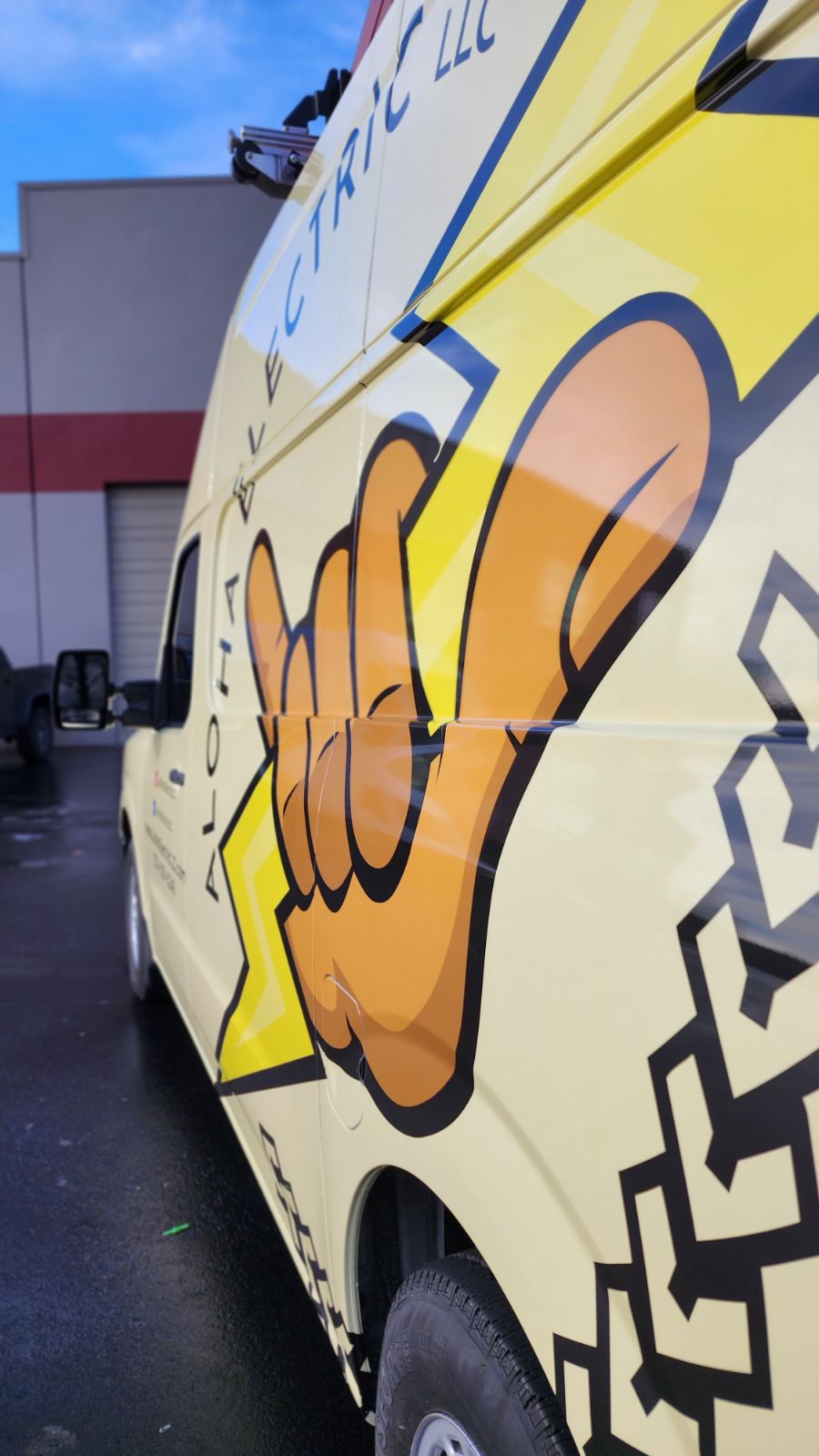 A yellow van with “Aloha Electric LLC” branding features a large shaka hand graphic on its side, parked outside a building with a red stripe.