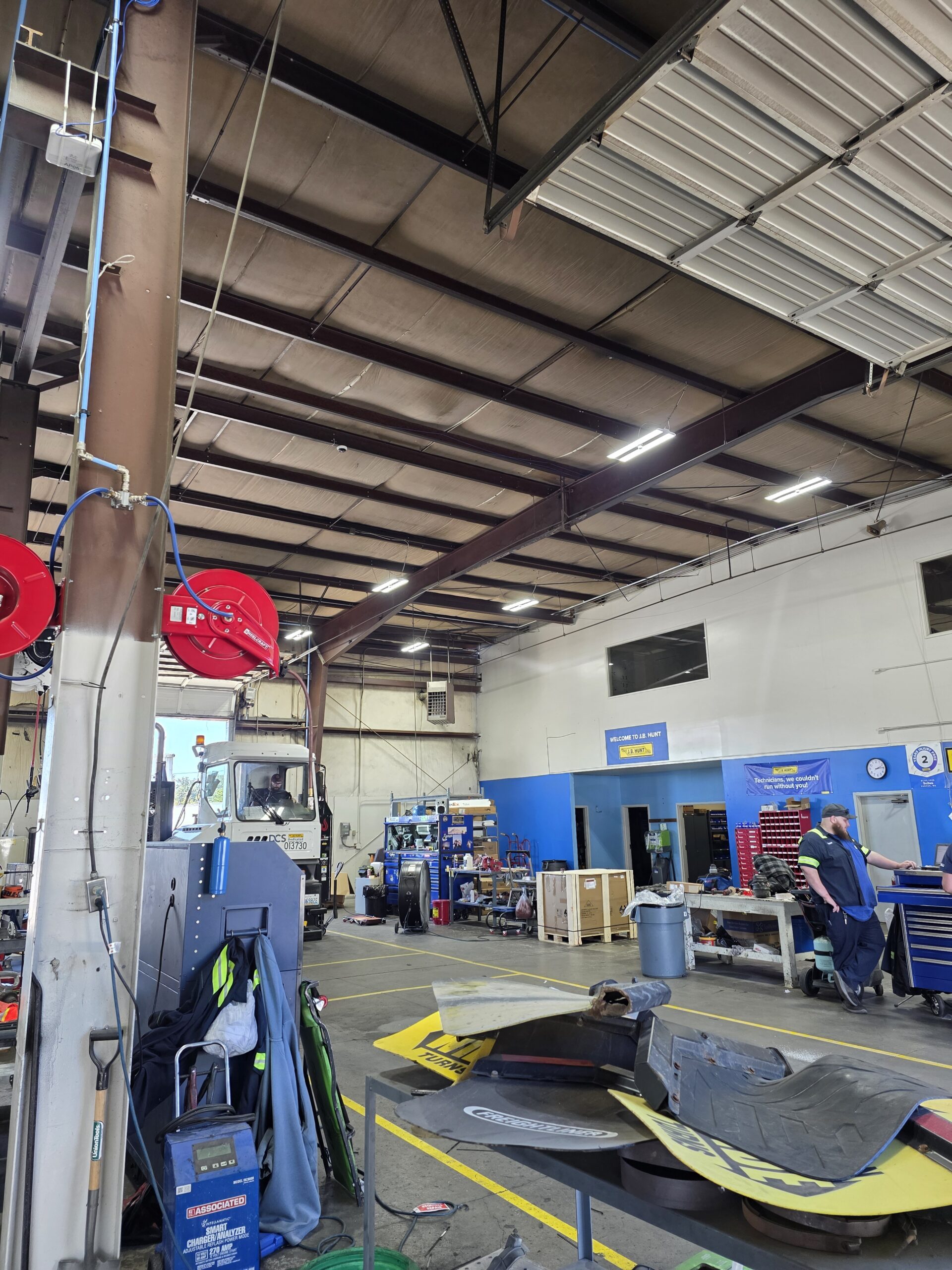 Interior of an auto repair shop with tools, machinery, and equipment scattered around; a worker stands near a blue wall in the background.