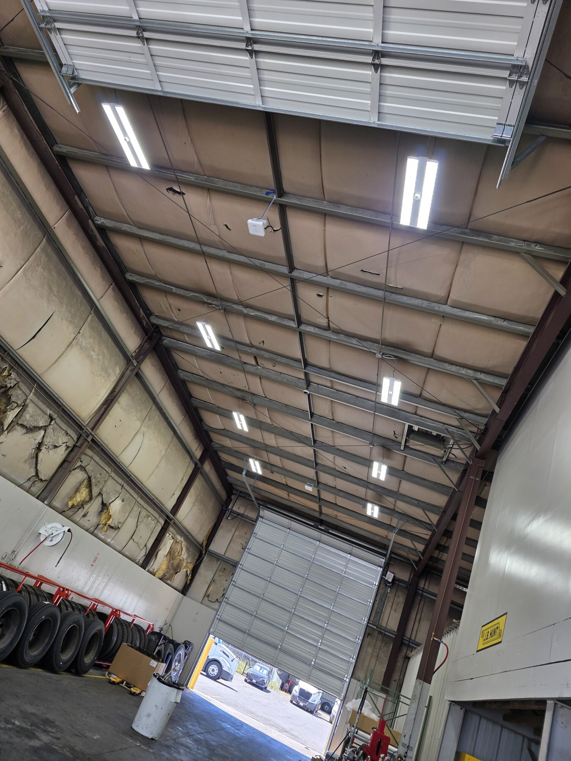 Interior of a large garage with an open overhead door, fluorescent ceiling lights, tires lined against one wall, and vehicles visible outside.