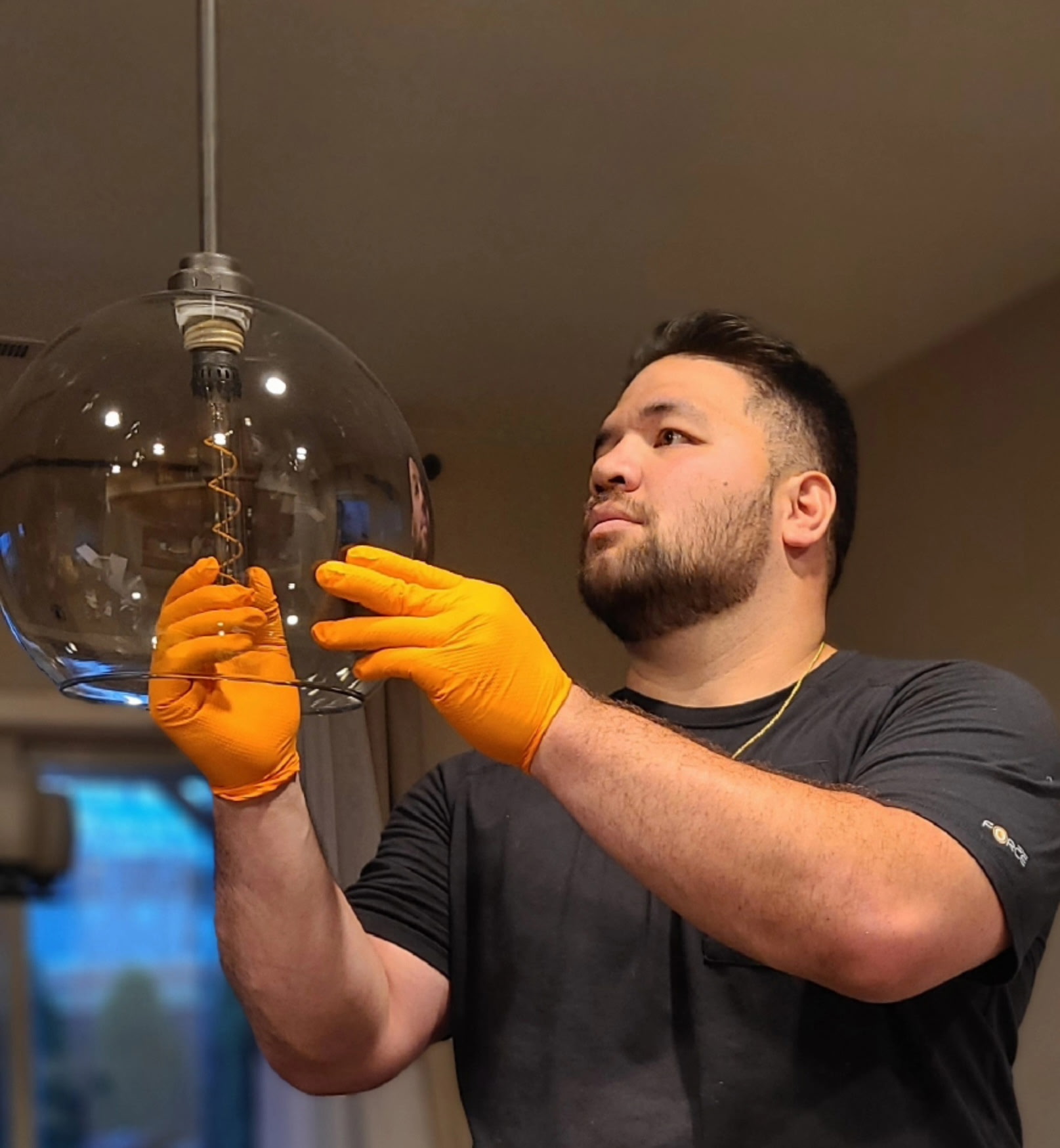 A man wearing orange gloves installs or cleans a clear glass light fixture indoors.